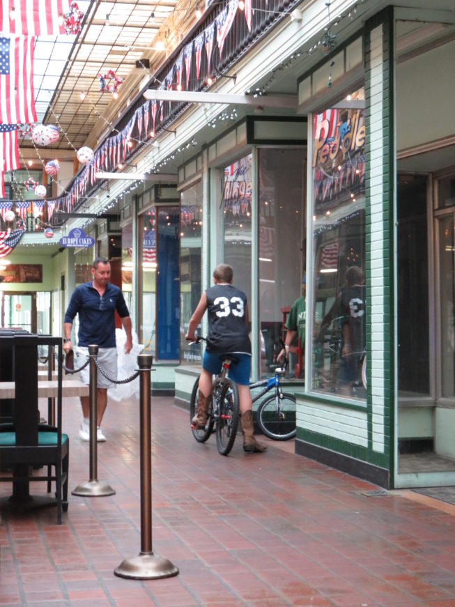 Robert Dalton, proprietor of the Paddock Club, walks through the delicious aromas from Europe Cakes as a visitor to the Arcade rides a bike around inside by Village Peddler, a bike and repair shop. Photo by Camilla Ammirati, 2017.